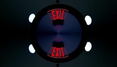 A symmetrical, kaleidoscope-like image featuring the word 'EXIT' in bold red letters repeated twice. The letters are displayed at opposite ends, surrounded by a dark circle with a gradient of blue and purple at the center. Bright circular lights appear at the four corners of the image, contributing to the geometric and symmetrical design.