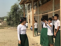 A group of school children in uniforms are gathered outside a building. Most of the children are standing close to each other and appear to be interacting with laughter and gestures. One girl stands slightly apart to the left, seemingly amused. The background features a brick building with wooden supports and a tree, indicating a rural or village setting.
