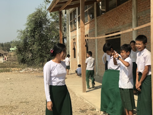 A group of school children in uniforms are gathered outside a building. Most of the children are standing close to each other and appear to be interacting with laughter and gestures. One girl stands slightly apart to the left, seemingly amused. The background features a brick building with wooden supports and a tree, indicating a rural or village setting.