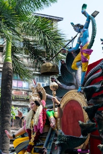 A vibrant statue of a deity adorned with colorful garments and floral decorations sits majestically on a platform. Another figure, painted blue, is depicted playing a flute, surrounded by ornate carvings and a multi-headed snake. The background showcases lush palm trees and a building.