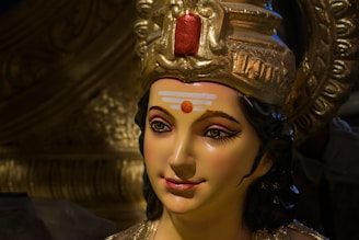 A detailed sculpture of a deity with a serene expression. The sculpture features intricate details in the crown, with gold accents and a prominent red element at the top. The face has carefully painted features, including long eyelashes and a calm smile, with a distinct tilak on the forehead.