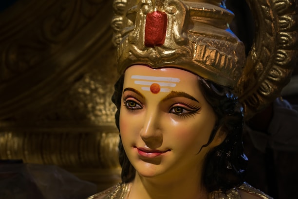 A detailed sculpture of a deity with a serene expression. The sculpture features intricate details in the crown, with gold accents and a prominent red element at the top. The face has carefully painted features, including long eyelashes and a calm smile, with a distinct tilak on the forehead.