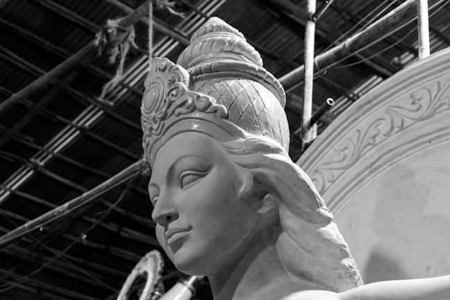 A detailed sculpture of a serene face with ornate headgear, likely in a workshop setting indicated by scaffolding and ropes in the background. The craftsmanship highlights intricate carvings and a smooth finish.
