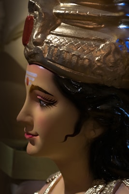 A detailed side profile of a sculpture with delicate facial features, intricate makeup, and a golden headdress adorned with elaborate designs. The sculpture appears to be a representation of a deity or historical figure.