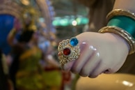 A close-up of a hand adorned with colorful jewelry, including rings featuring red and blue stones set in ornate gold designs. The wrist is decorated with green and gold bangles, and the background is blurred with an impression of vibrant colors and patterns.