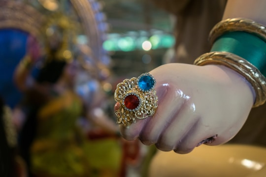 A close-up of a hand adorned with colorful jewelry, including rings featuring red and blue stones set in ornate gold designs. The wrist is decorated with green and gold bangles, and the background is blurred with an impression of vibrant colors and patterns.