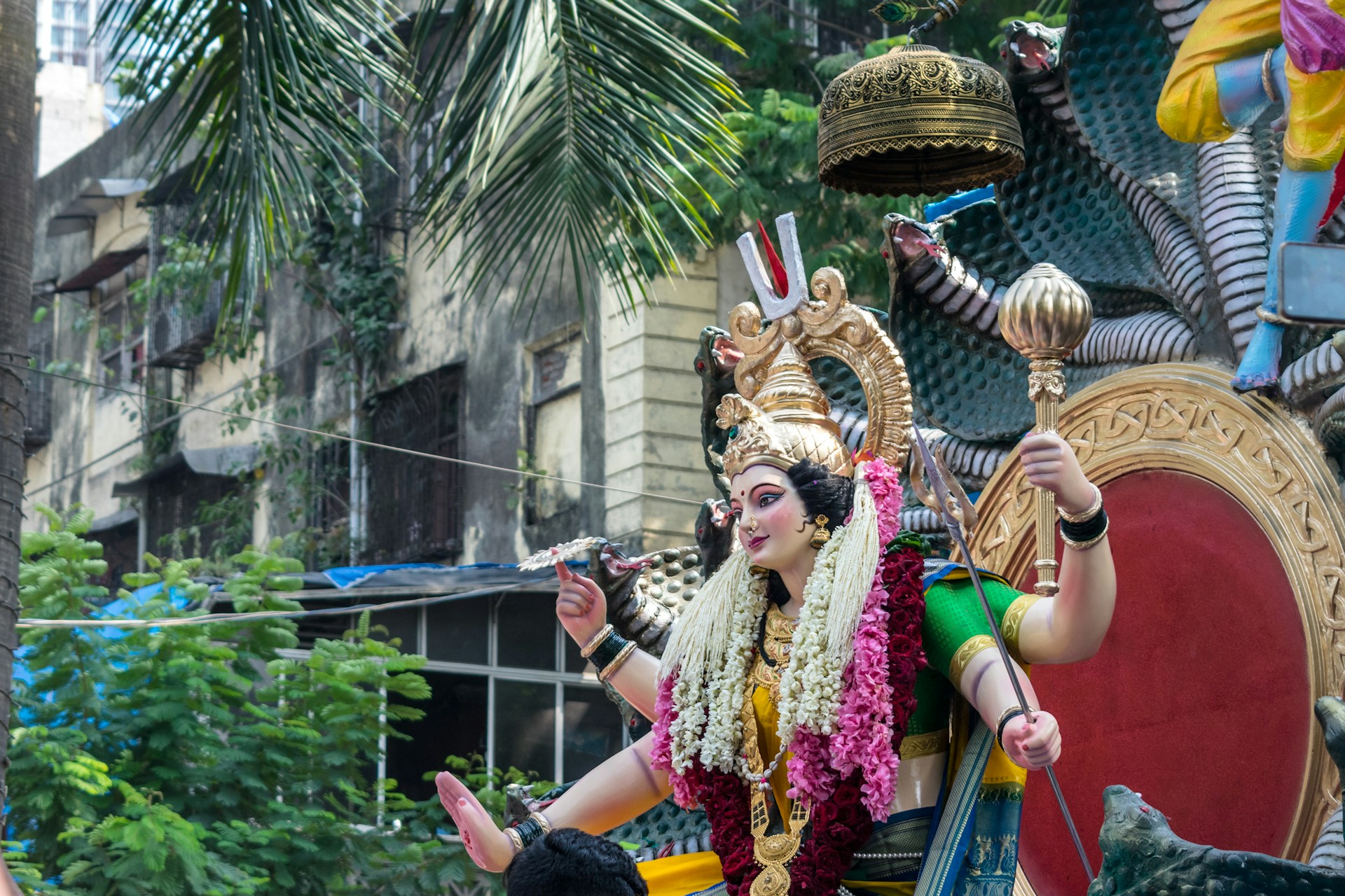 A vibrant statue of a deity adorned with intricate jewelry, floral garlands, and traditional attire. The statue is in a dynamic pose, holding a scepter and a serpent, with a large decorative headdress. The background features urban architectural elements with palm leaves providing a natural contrast.