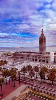 A waterfront scene featuring a historic clock tower with scaffolding, palm trees lining a broad street, and a large building with arched windows. In the background, a body of water stretches out to the horizon where a cargo ship can be seen, under a sky filled with scattered clouds.