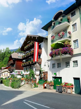 Charming alpine village with traditional chalet-style buildings featuring intricate wooden balconies adorned with vibrant flower boxes. An Austrian flag is prominently displayed, contrasting with the lush greenery and blue sky.