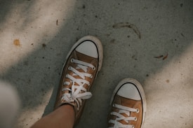 A pair of brown canvas sneakers with white laces and rubber caps are worn by a person standing on a rough, slightly dirty concrete surface. The sneakers appear to be well-worn, and there are shadows cast on the ground, adding a textured look to the scene.