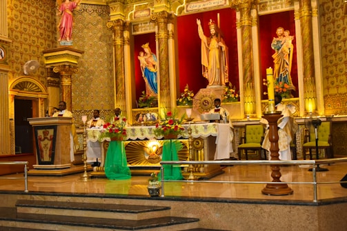 A richly decorated church altar with ornate columns and religious statues. Clergymen, dressed in ceremonial robes, conduct a service. The altar is adorned with vibrant flowers and lit candles, set against a backdrop of intricate patterns and warm lighting.