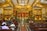 A richly decorated interior of a church featuring a grand altar with religious statues and ornate golden designs. Stained glass windows, intricate ceiling patterns, and a congregation seated in wooden pews add to the ambiance.