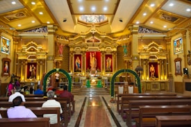 A richly decorated interior of a church featuring a grand altar with religious statues and ornate golden designs. Stained glass windows, intricate ceiling patterns, and a congregation seated in wooden pews add to the ambiance.