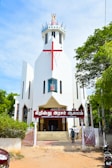 A modern white church with a large red cross on its facade. The building features a unique, angular design with a crown-like structure at the top. The entrance is adorned with a sign in a local script, and steps lead up to double doors. The surrounding area includes lush green trees and a clear blue sky, adding vibrant colors to the scene.