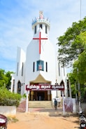 A modern white church with a large red cross on its facade. The building features a unique, angular design with a crown-like structure at the top. The entrance is adorned with a sign in a local script, and steps lead up to double doors. The surrounding area includes lush green trees and a clear blue sky, adding vibrant colors to the scene.