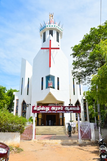 A modern white church with a large red cross on its facade. The building features a unique, angular design with a crown-like structure at the top. The entrance is adorned with a sign in a local script, and steps lead up to double doors. The surrounding area includes lush green trees and a clear blue sky, adding vibrant colors to the scene.