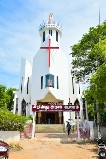 A modern white church with a large red cross on its facade. The building features a unique, angular design with a crown-like structure at the top. The entrance is adorned with a sign in a local script, and steps lead up to double doors. The surrounding area includes lush green trees and a clear blue sky, adding vibrant colors to the scene.