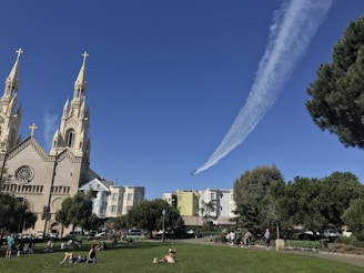 In a vibrant park setting, groups of people are relaxing on the grass. A large church with two tall, ornate steeples and gold crosses stands prominently on the left. In the sky, an airplane leaves a long, dramatic trail of white smoke, curving gracefully across the bright blue sky.