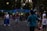 A person is creating a large, colorful bubble in a park setting. Several people are gathered around, observing the activity with interest, including a young girl holding a balloon animal. The background is filled with trees and a pathway, with street lamps faintly illuminating the scene as dusk approaches.