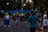 A person is creating a large, colorful bubble in a park setting. Several people are gathered around, observing the activity with interest, including a young girl holding a balloon animal. The background is filled with trees and a pathway, with street lamps faintly illuminating the scene as dusk approaches.