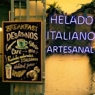 A chalkboard menu lists various breakfast items and beverages like cappuccino and espresso, alongside natural and Italian products. Next to it, glowing neon signs display the words 'HELADO ITALIANO ARTESANAL' against a background adorned with green plants.