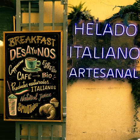 A chalkboard menu lists various breakfast items and beverages like cappuccino and espresso, alongside natural and Italian products. Next to it, glowing neon signs display the words 'HELADO ITALIANO ARTESANAL' against a background adorned with green plants.