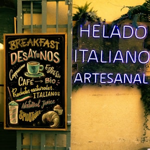 A chalkboard menu lists various breakfast items and beverages like cappuccino and espresso, alongside natural and Italian products. Next to it, glowing neon signs display the words 'HELADO ITALIANO ARTESANAL' against a background adorned with green plants.