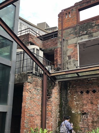 A partially dilapidated brick building features exposed red bricks and overgrown vegetation climbing its walls. Metal beams frame some parts of the structure, hinting at a modern renovation. A person appears to be observing or photographing the site, adding a sense of curiosity and exploration. The overall look of the construction is rustic and industrial with visible wear and decay on the bricks.