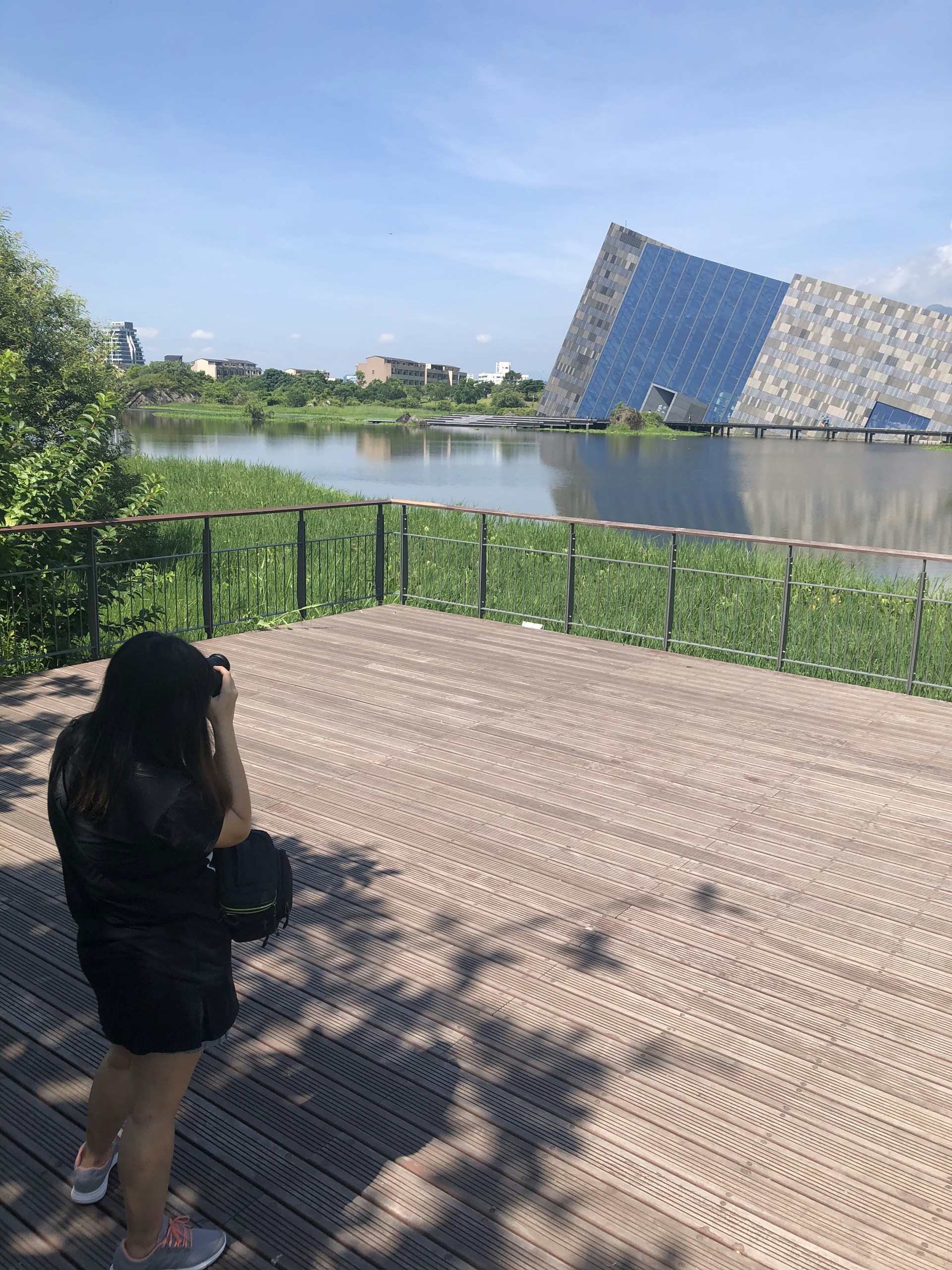 A person is standing on a wooden deck taking a photograph of a modern, angular building across a body of water. The building has a unique tilted design with blue and gray panels. The surrounding area is lush with green vegetation.