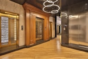 An elegant elevator lobby with three elevators, two with ornate wood paneling and one with a modern metallic finish. A stylish light fixture with circular designs hangs overhead, and the floor is covered with polished wooden planks.