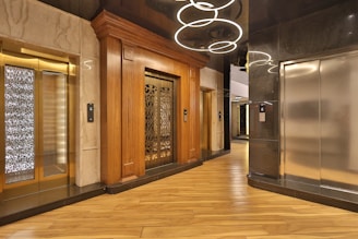 An elegant elevator lobby with three elevators, two with ornate wood paneling and one with a modern metallic finish. A stylish light fixture with circular designs hangs overhead, and the floor is covered with polished wooden planks.