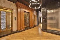 An elegant elevator lobby with three elevators, two with ornate wood paneling and one with a modern metallic finish. A stylish light fixture with circular designs hangs overhead, and the floor is covered with polished wooden planks.