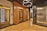 An elegant elevator lobby with three elevators, two with ornate wood paneling and one with a modern metallic finish. A stylish light fixture with circular designs hangs overhead, and the floor is covered with polished wooden planks.