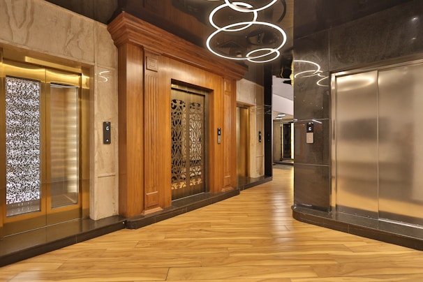 An elegant elevator lobby with three elevators, two with ornate wood paneling and one with a modern metallic finish. A stylish light fixture with circular designs hangs overhead, and the floor is covered with polished wooden planks.