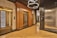 An elegant elevator lobby with three elevators, two with ornate wood paneling and one with a modern metallic finish. A stylish light fixture with circular designs hangs overhead, and the floor is covered with polished wooden planks.