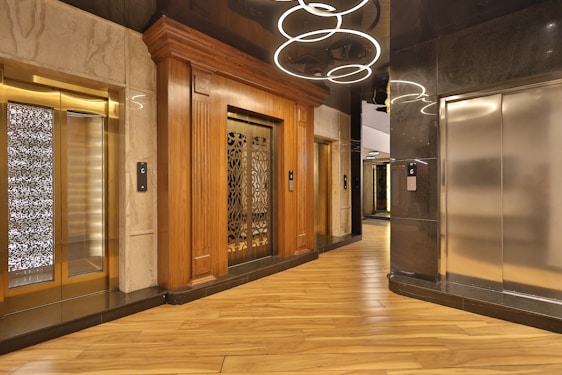 An elegant elevator lobby with three elevators, two with ornate wood paneling and one with a modern metallic finish. A stylish light fixture with circular designs hangs overhead, and the floor is covered with polished wooden planks.