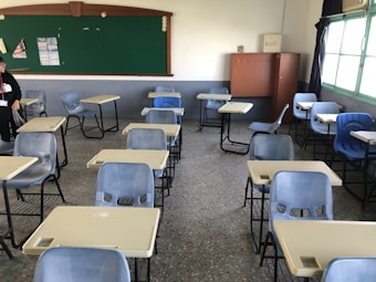 A classroom with several empty blue and beige desks arranged in rows. A person is standing near the back of the room, wearing a black hoodie. The chalkboard at the front is largely unused, with a few posters or papers pinned to it. The room has terrazzo flooring, and there is a large window with light streaming in, casting natural light across the room. A wooden cabinet is positioned near the back wall.