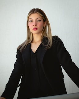 A woman with straight blonde hair, wearing a black suit jacket over a black top. She is standing against a plain, light-colored background and has a serious or contemplative expression on her face. Her makeup is done in a subtle yet elegant style with bold eyeliner and red lipstick.