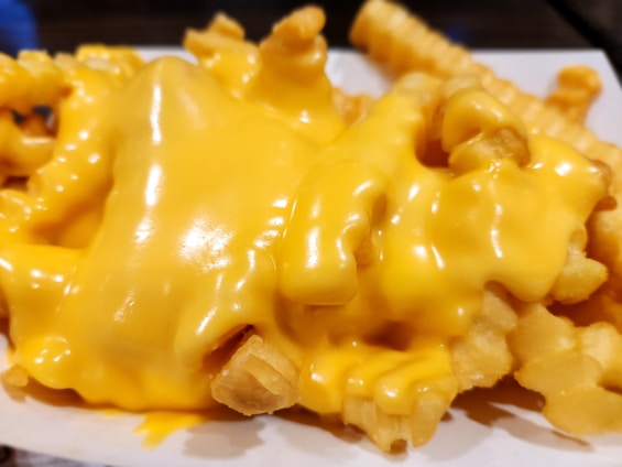 A close-up view of crinkle-cut french fries covered in melted cheese sauce, creating a rich and creamy appearance. The cheese glistens under the light, emphasizing its smooth texture and vibrant color.