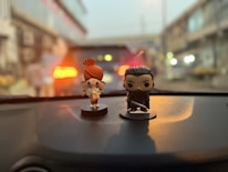 Two small figurines are placed on a car dashboard. The one on the left is a colorful character with large eyes, a big red hair bun, and a playful outfit. The figure on the right is a darker, more serious character holding a sword. The background is blurred, suggesting motion or focus on the figures.