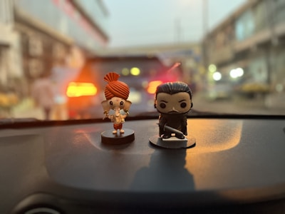 Two small figurines are placed on a car dashboard. The one on the left is a colorful character with large eyes, a big red hair bun, and a playful outfit. The figure on the right is a darker, more serious character holding a sword. The background is blurred, suggesting motion or focus on the figures.