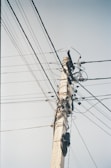 A tall utility pole stands against a clear sky, with an intricate network of cables and wires extending in various directions. The pole is made of concrete, and multiple insulators and clamps are visible connecting the wires. The image captures the complexity of modern infrastructure with numerous power and communication lines intertwined.