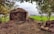 A rustic hut made of earth and straw is situated in a rural setting. The foreground is filled with dry brown soil and some scattered branches. Lush green fields stretch out towards the horizon, bordered by rows of trees. The scene is framed by trees with dense foliage on both sides.