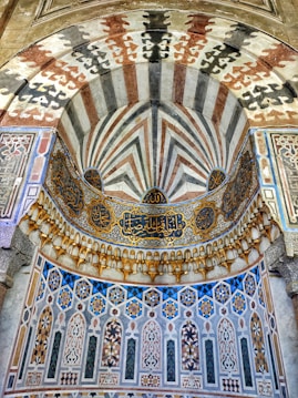 Intricate Islamic architectural design featuring ornate geometric patterns and Arabic calligraphy. The structure is adorned with colorful mosaic tiles and showcases an array of arches with a combination of pointed and horseshoe shapes.