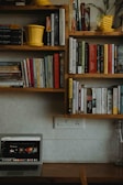 A wooden bookshelf filled with various books is displayed on a light-colored wall. Two yellow planters are placed on the shelves, adding a pop of color. Below the shelves, a desk holds a laptop with a website open on the screen displaying various content. A glass bottle is positioned on the right side of the desk, near an electrical outlet.