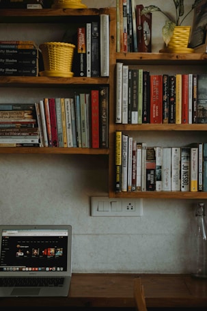 A wooden bookshelf filled with various books is displayed on a light-colored wall. Two yellow planters are placed on the shelves, adding a pop of color. Below the shelves, a desk holds a laptop with a website open on the screen displaying various content. A glass bottle is positioned on the right side of the desk, near an electrical outlet.