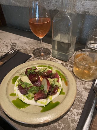A gourmet dish is beautifully presented on a rustic stoneware plate. It features a delicate arrangement of thinly sliced food, garnished with fresh herbs and drizzled with green sauce. Beside the plate, there is a glass of rosé wine, a glass of whisky with ice, and a clear water bottle, placed on a stone surface.