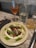 A gourmet dish is beautifully presented on a rustic stoneware plate. It features a delicate arrangement of thinly sliced food, garnished with fresh herbs and drizzled with green sauce. Beside the plate, there is a glass of rosé wine, a glass of whisky with ice, and a clear water bottle, placed on a stone surface.