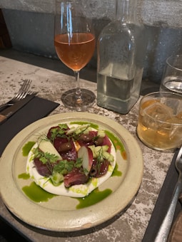 A gourmet dish is beautifully presented on a rustic stoneware plate. It features a delicate arrangement of thinly sliced food, garnished with fresh herbs and drizzled with green sauce. Beside the plate, there is a glass of rosé wine, a glass of whisky with ice, and a clear water bottle, placed on a stone surface.