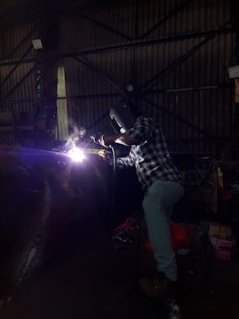 A person wearing a welding helmet is working on a metal structure with a bright welding torch. The surroundings are dark, with industrial equipment and a corrugated metal wall visible in the background.